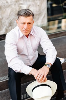 Serious man sitting on pavement and holding a hat in his hands, looking at the camera. - Image photo