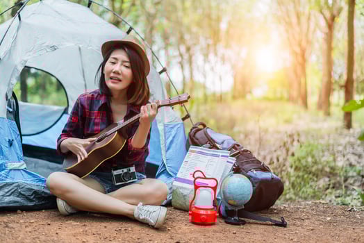 Beautiful Asian woman playing Ukulele in front of camping tent in pine woods. People and Lifestyles concept. Adventure and Travel theme photo