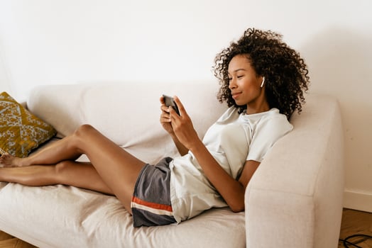 Black young woman wearing earphones using mobile phone while resting on sofa photo