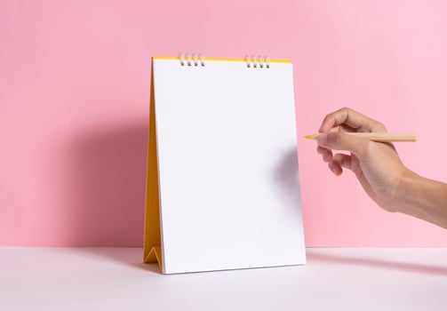 Hand holding pencil for writing marking on mockup paper spiral calendar on pink background. photo