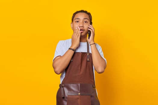 Shocked asian man covering mouth with hands and holding smartphone over yellow background photo
