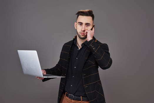 Busy bearded man in business clothes talking by smartphone and using laptop computer while holding cup of coffee in hand over gray background photo