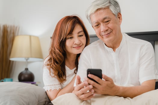 Asian senior couple using mobile phone at home. Asian Senior Chinese grandparents, husband and wife happy after wake up, watching movie lying on bed in bedroom at home in the morning concept. photo