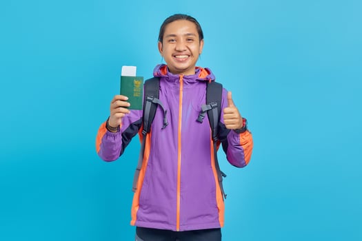 Portrait of cheerful Asian young man showing passport book and giving thumbs up isolated on blue background photo