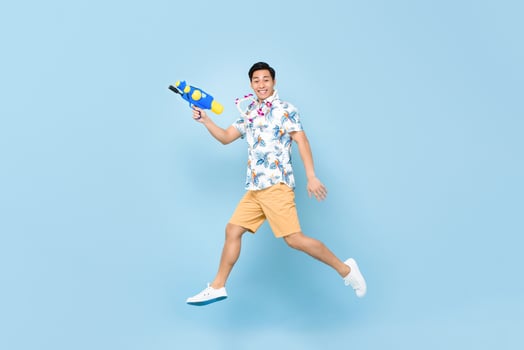 Handsome smiling young Asian man playing with water gun and jumping in studio blue background for Songkran festival in Thailand and southeast Asia photo