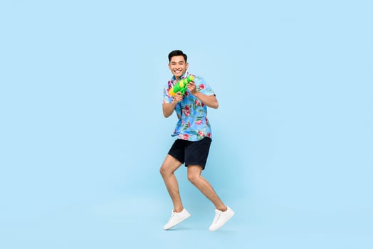 Handsome smiling young Asian man playing with water gun and jumping in studio blue background for Songkran festival in Thailand and southeast Asia photo