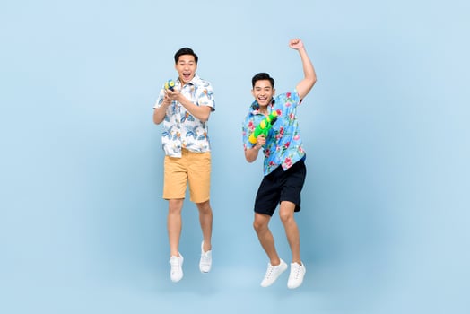 Smilng happy Asian male friends playing with water guns and jumping in blue isolated background for Songkran festival in Thailand and southeast Asia photo