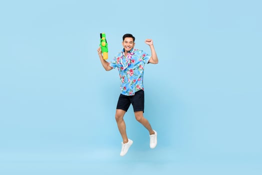 Handsome smiling young Asian man playing with water gun and jumping in studio blue background for Songkran festival in Thailand and southeast Asia photo