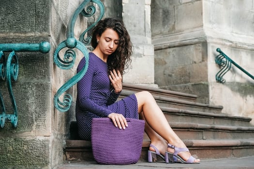 Woman wearing purple polka dot dress, holding knitted bag sitting on the stairs posing in street of city. Copy, empty space for text photo