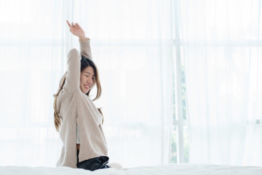 Happy beautiful young Asian woman waking up in morning, sitting on bed, stretching in cozy bedroom, looking through window. Funny asian woman after wake up. Asia woman is stretching and smiling. photo