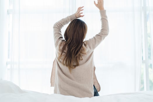 Back view of happy beautiful young Asian woman waking up in morning, sitting on bed, stretching in cozy bedroom, looking through window. Funny woman after wake up. She is stretching and smiling. photo