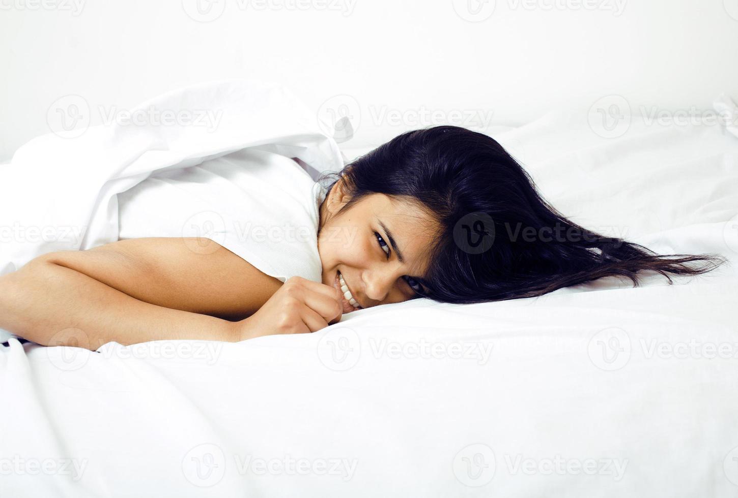 mujer real morena bastante india en la cama sonriendo, sábanas blancas foto