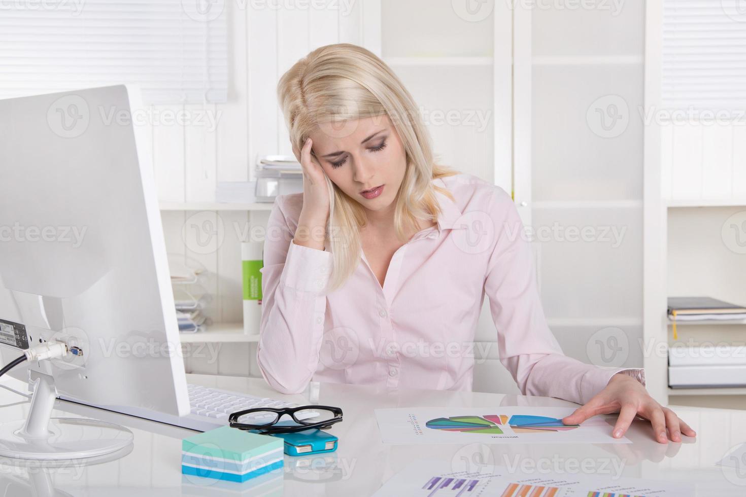 Young blonde trainee in rose blouse with headache at desk. photo