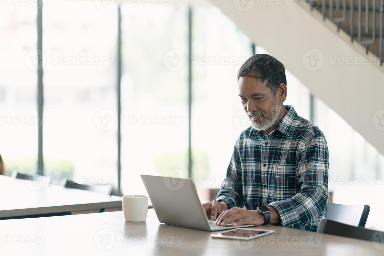 sonriente hombre maduro atractivo con blanco, gris elegante barba corta usando un teléfono inteligente que sirve internet en la oficina moderna, espacio de trabajo compartido o cafetería. Viejo usando tecnología de redes sociales. foto