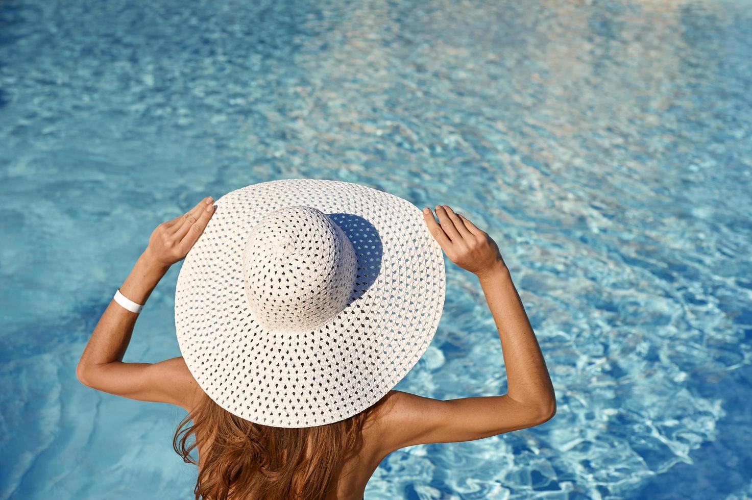 Vista trasera de la mujer con sombrero blanco sentada junto a la piscina en un día soleado. concepto de viaje por mar con lugar para el texto foto