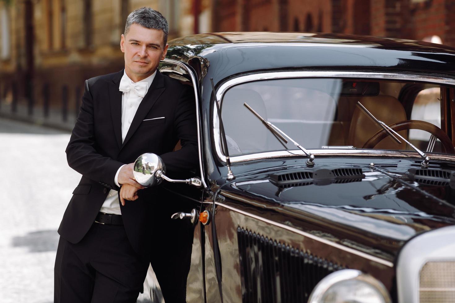 Handsome groom in black tuxedo poses before a black retro car photo