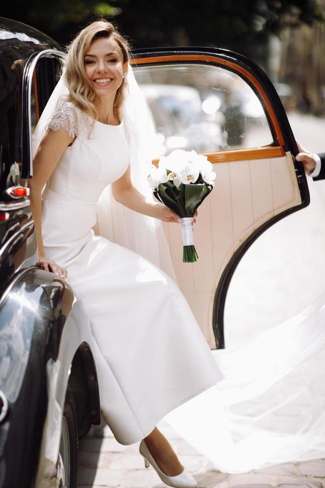 hermosa novia en vestido blanco de lujo sale de un coche retro foto