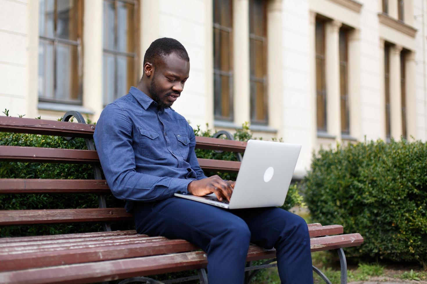 Happy African American man works on his laptop sitting on the bench outside photo