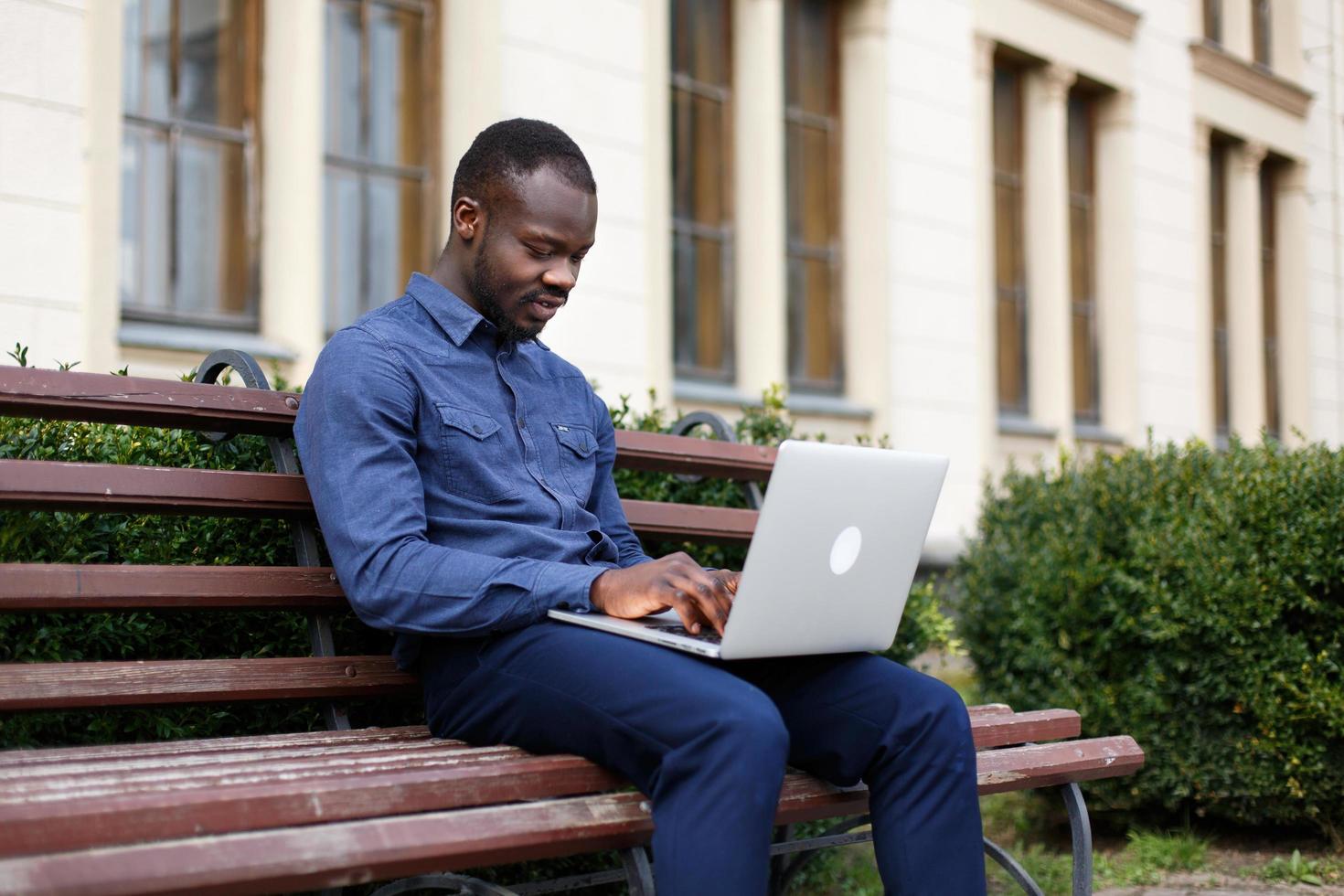 Feliz hombre afroamericano trabaja en su computadora portátil sentado en el banco fuera foto