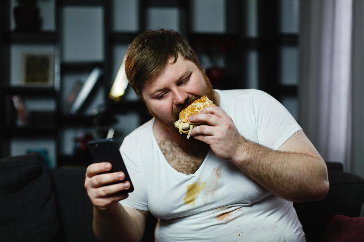 El hombre revisa algo en su teléfono inteligente mientras come una hamburguesa foto