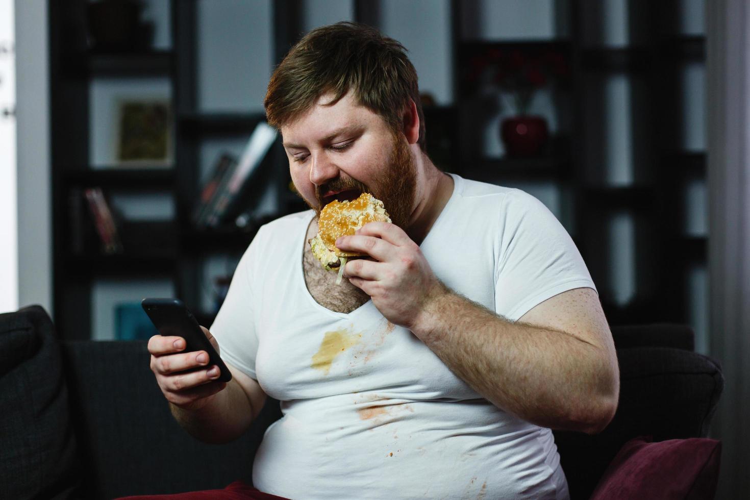 hombre gordo revisa algo en su teléfono inteligente mientras come una hamburguesa foto