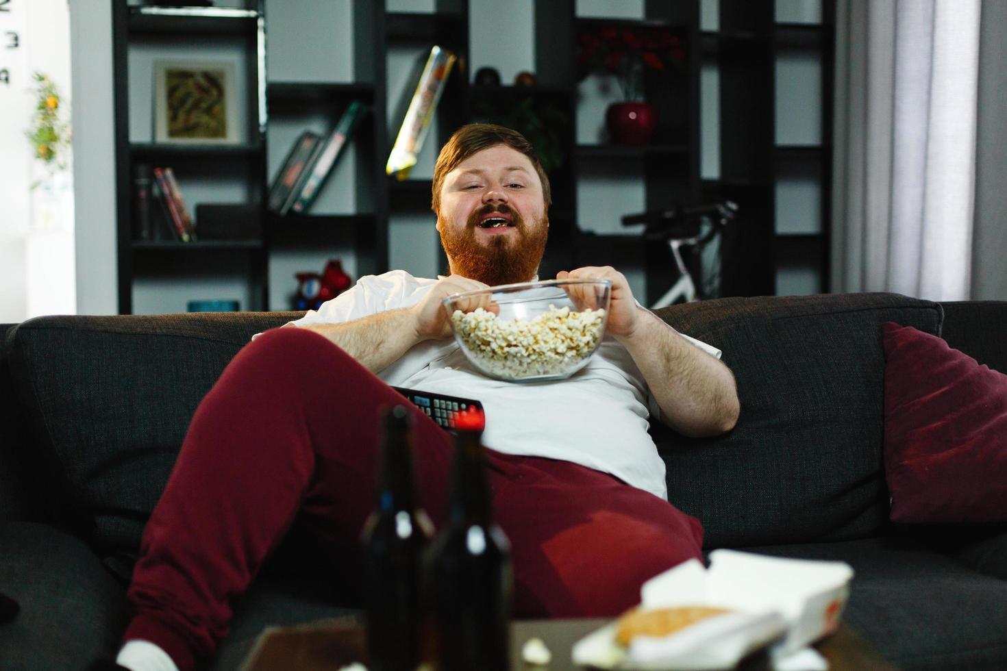 Hombre gordo feliz come palomitas de maíz acostado en el sofá ante una mesa con cerveza foto