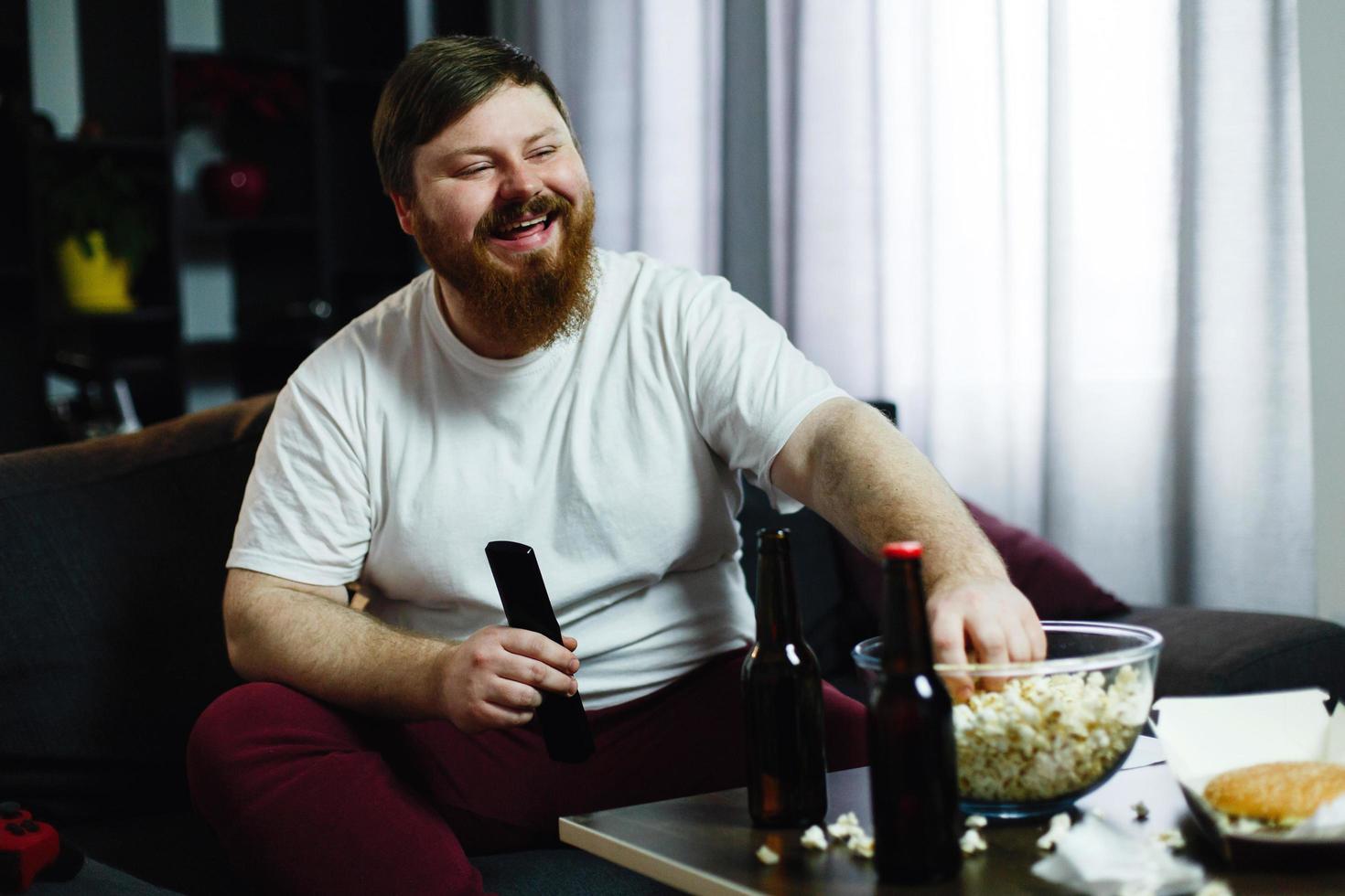 Happy fat man sits on the sofa and watches TV with popcorn and beer photo
