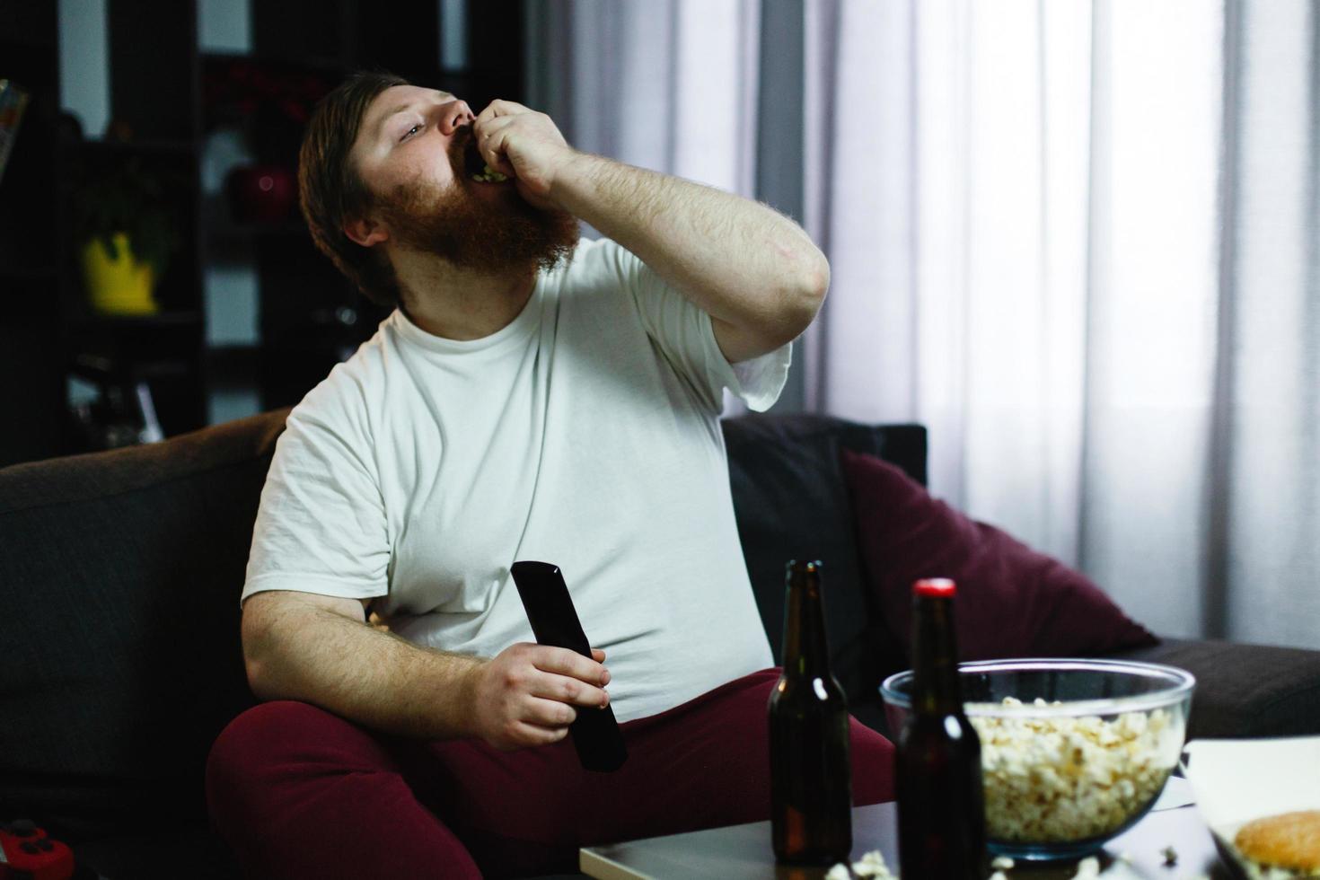 Hombre gordo feliz come palomitas de maíz acostado en el sofá ante una mesa con cerveza foto