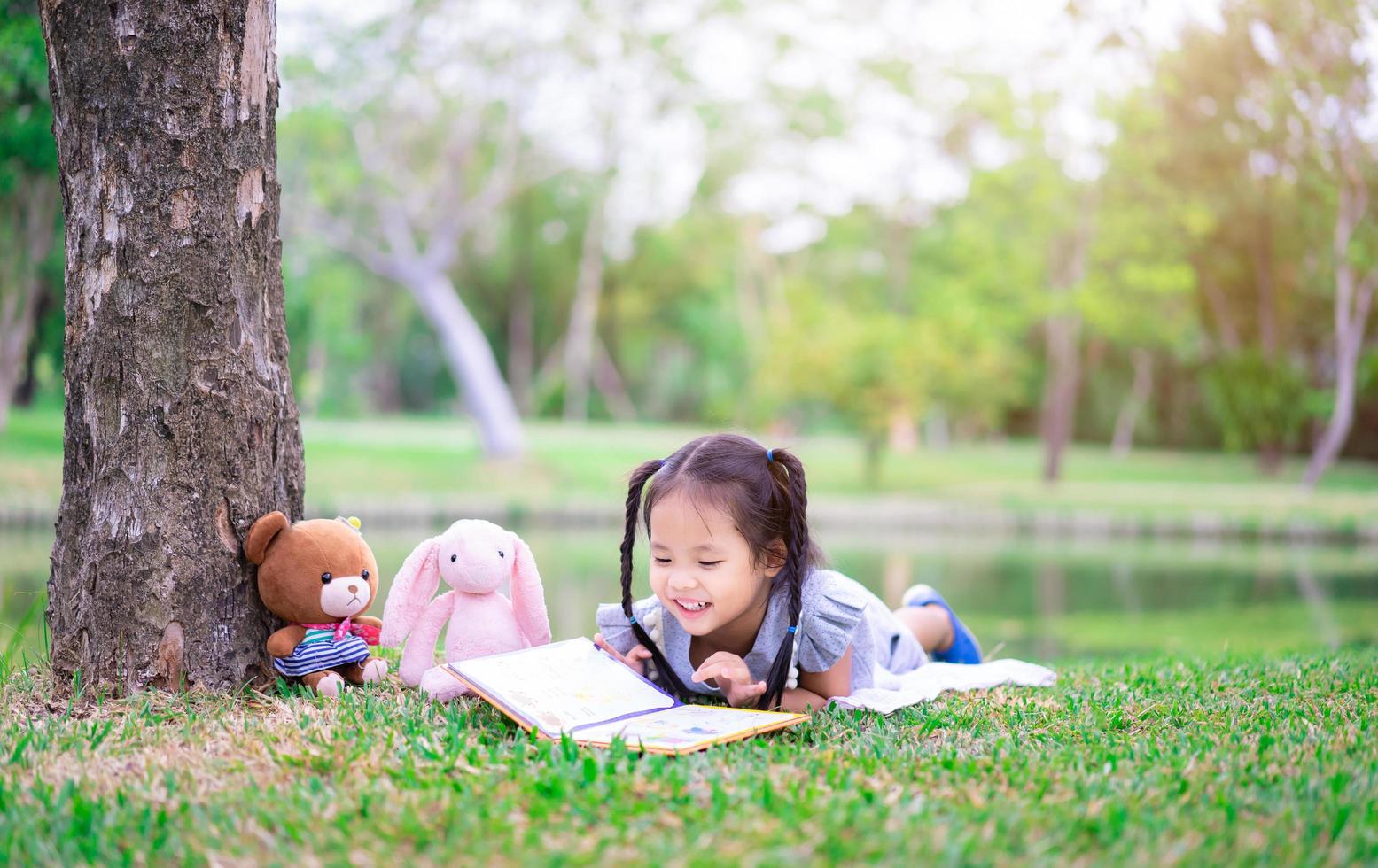 linda niña leyendo un libro mientras está acostada con una muñeca en el parque foto