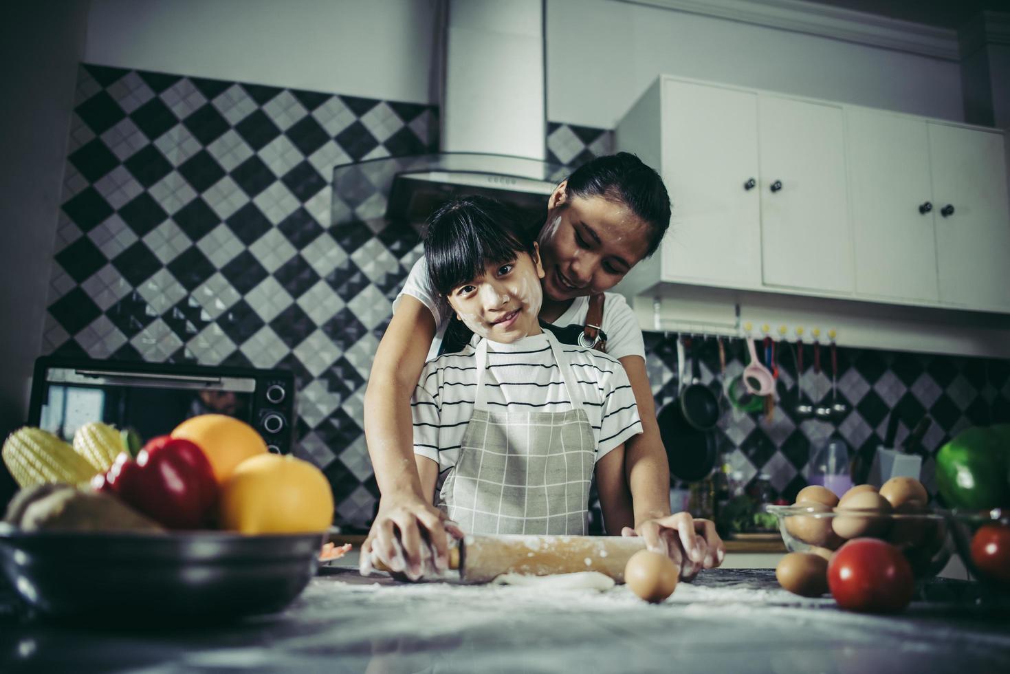 Cute little girl and mom in aprons flattening dough in home kitchen photo