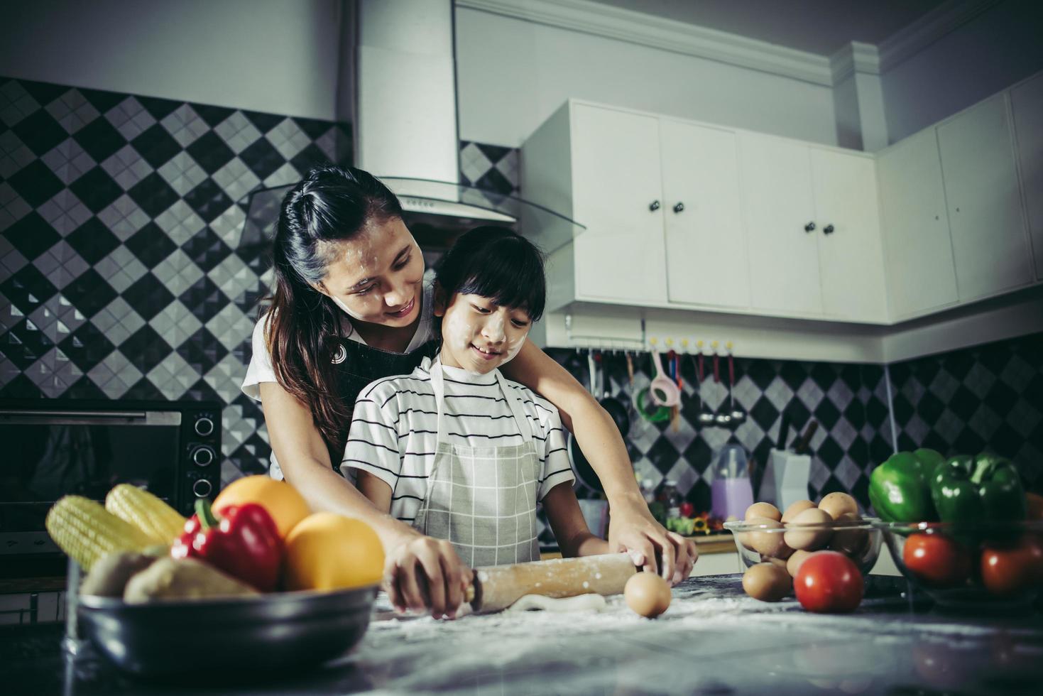 Linda niña y mamá en delantales aplanar la masa en la cocina de casa foto