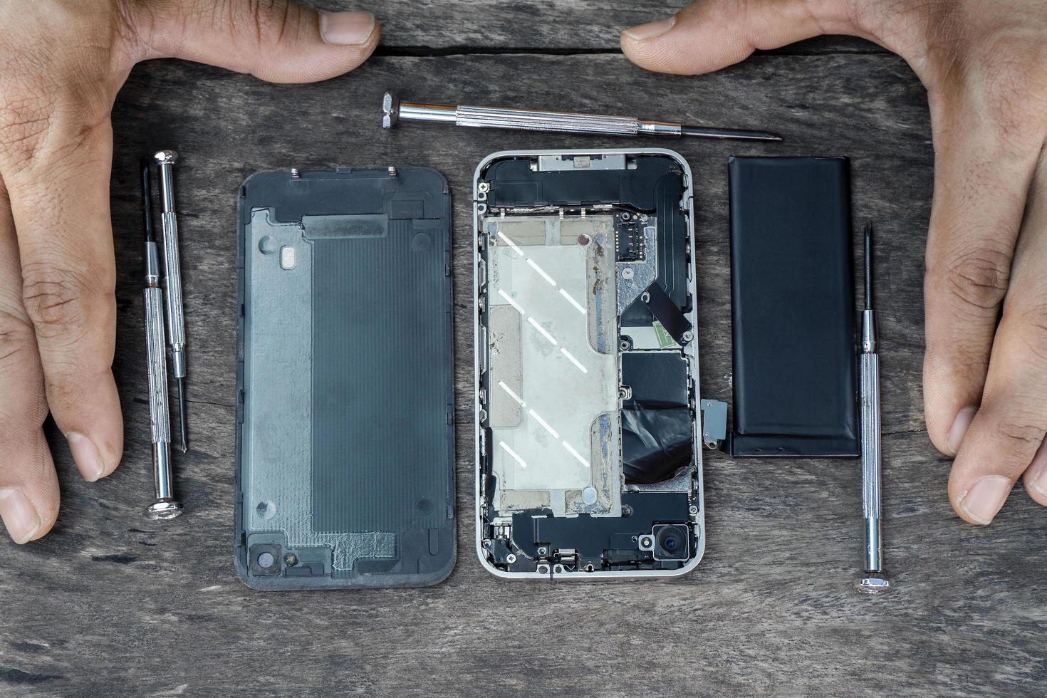 Top view of the hand technician with a screwdriver mobile phone repair inside a cell phone with a fixing battery from a broken service shop center on a wood table. Smartphone maintenance concept. photo