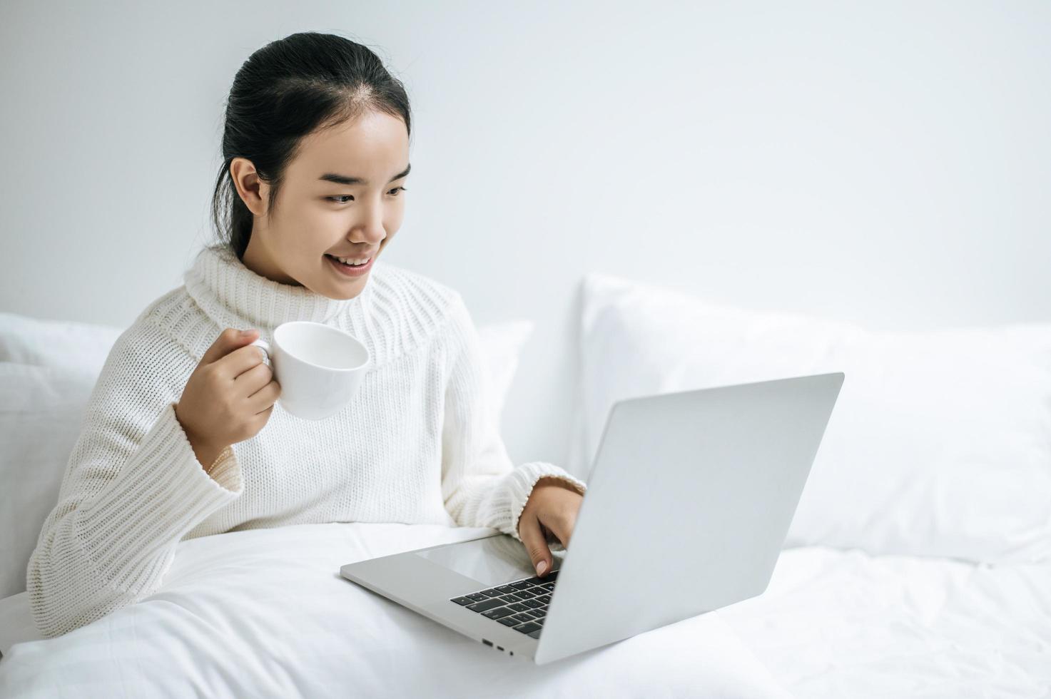 Mujer joven jugando en la computadora portátil y sosteniendo una taza de café en la cama foto