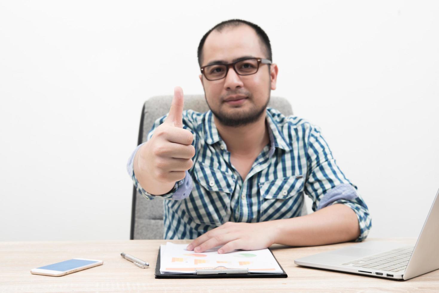 Retrato de joven empresario con Thumbs up sentado en el lugar de trabajo foto