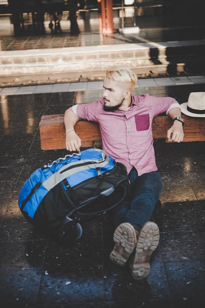 Hombre joven inconformista sentado en un banco de madera en la estación de tren foto