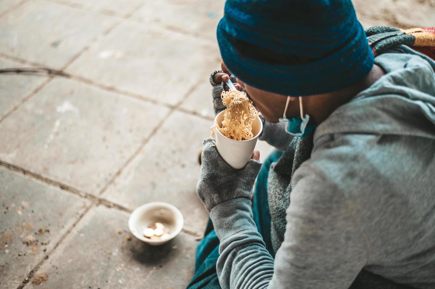A homeless man sits wrapped in cloth eating noodles photo