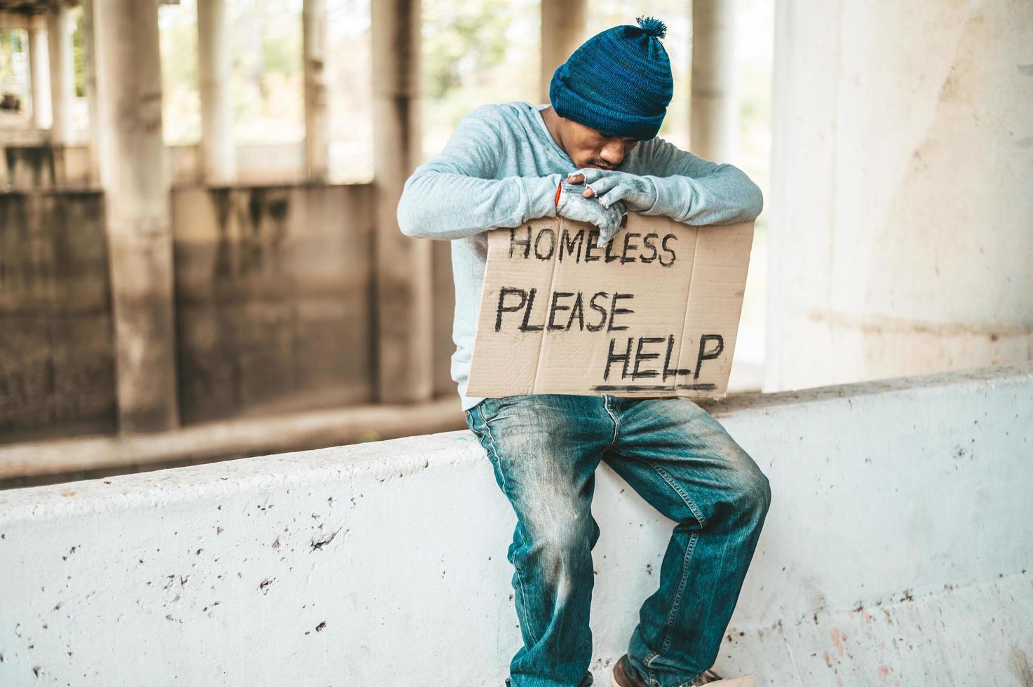 Beggar sits on a road barrier with homeless please help sign photo