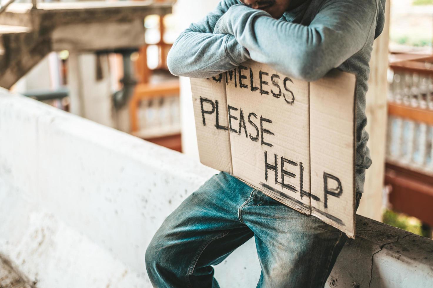 Beggar sits on a road barrier with homeless please help sign photo