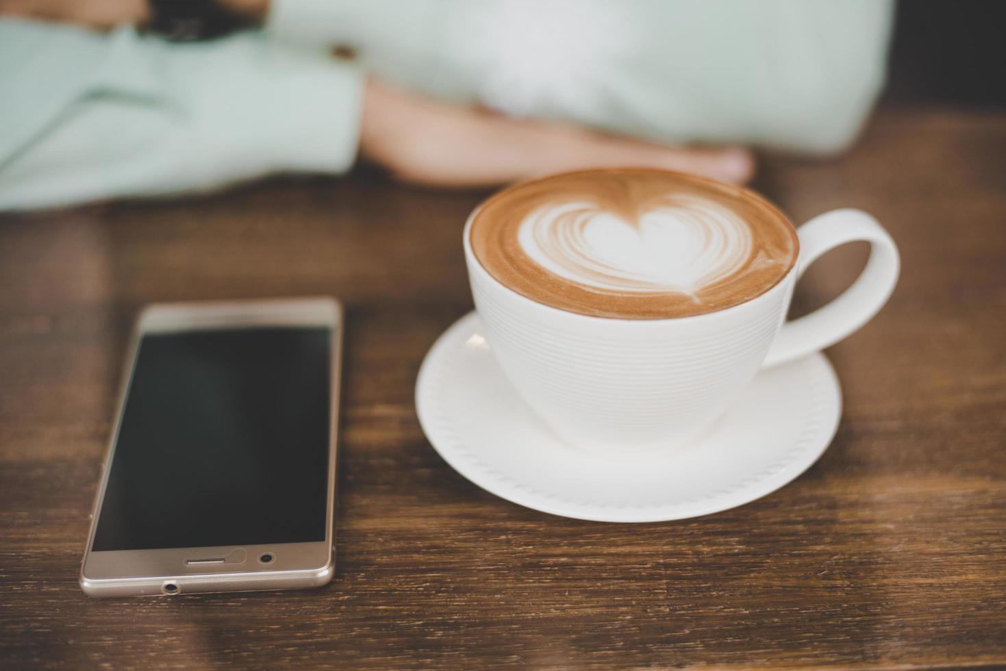 La mano del hombre con una taza de café con leche y un teléfono inteligente en el bar cafetería foto