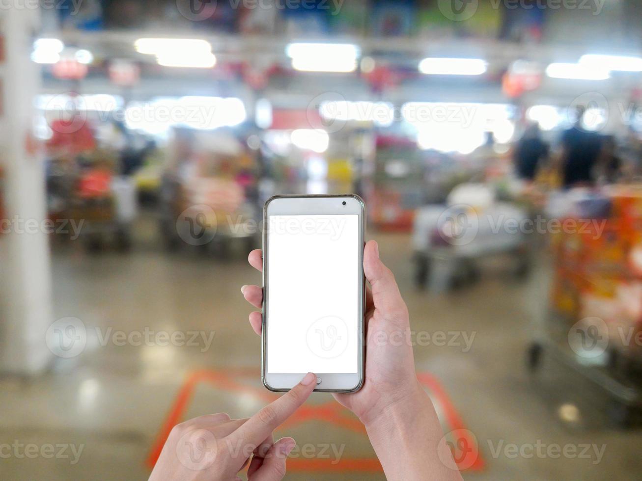 Mano con teléfono inteligente móvil de pantalla en blanco con fondo borroso en el supermercado foto