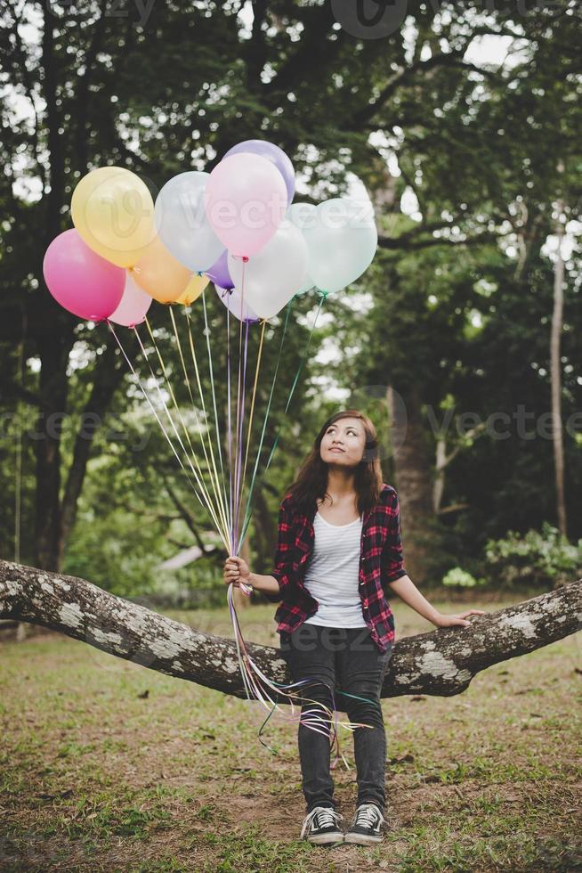 Young hipster woman sitting on tree branch with colorful balloons photo