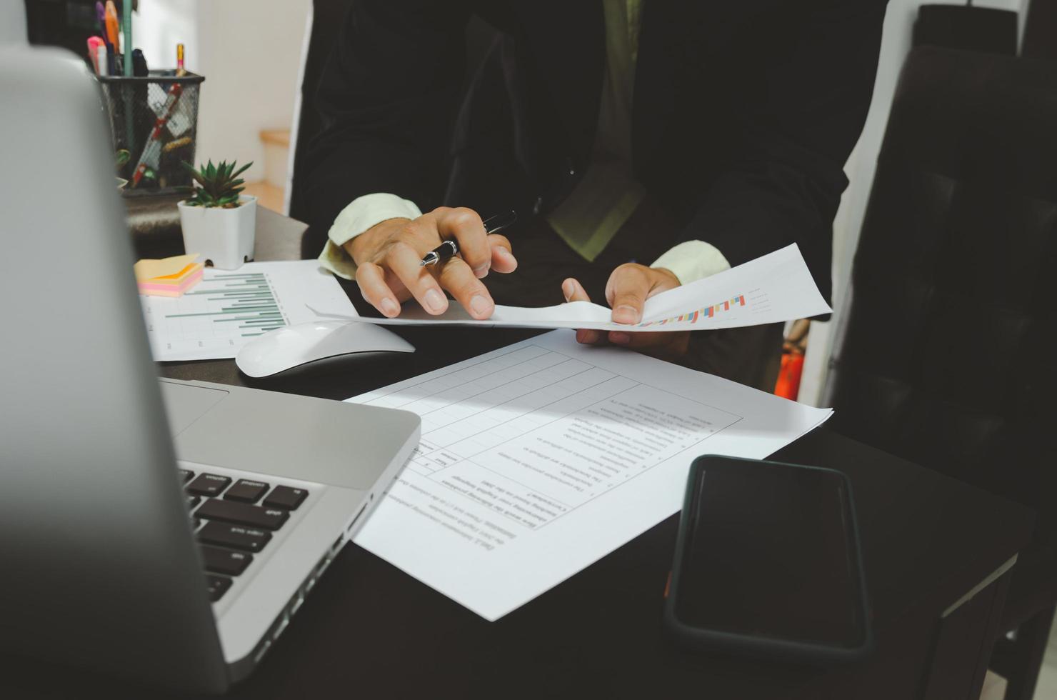 A businessman looking at business documents and holding a pen with a computer laptop and smartphone at the desk working from home photo