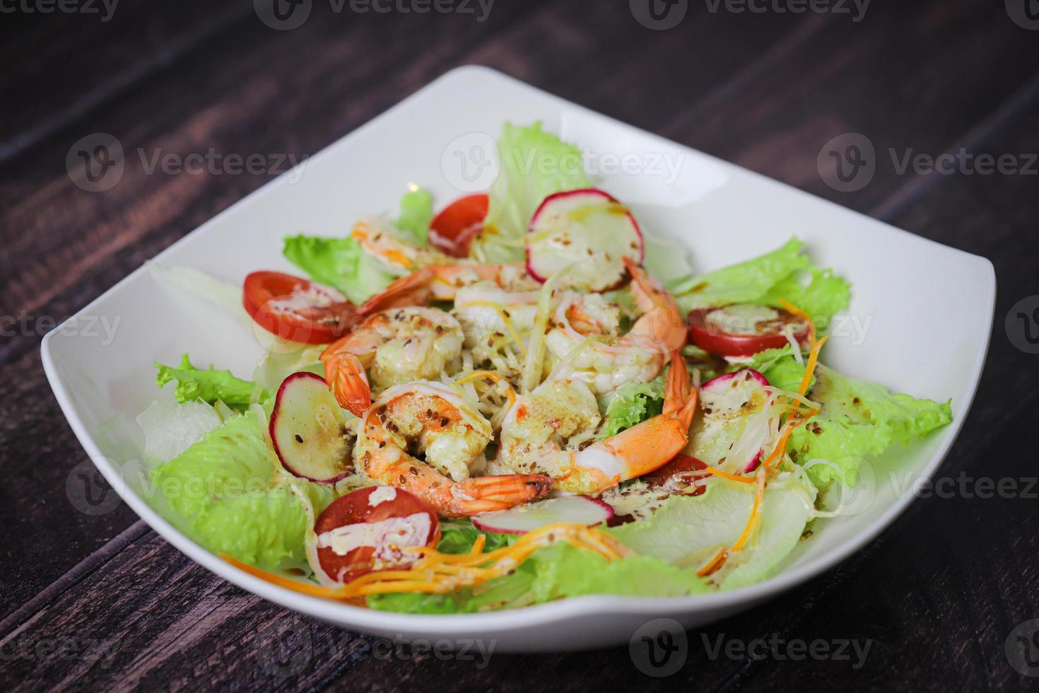 Ensalada con lechuga, rábanos, tomates cherry y camarones en plato blanco sobre mesa de madera foto