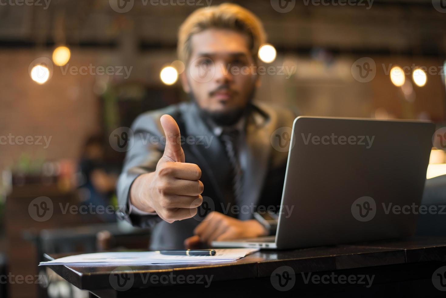 El empresario da Thumbs up mientras trabaja con un ordenador portátil en la cafetería. foto