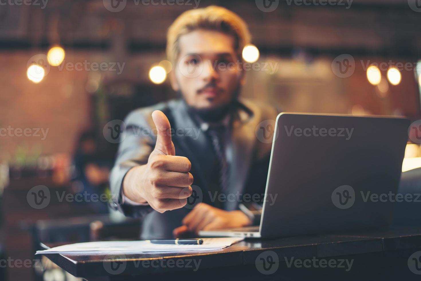 El empresario da Thumbs up mientras trabaja con un ordenador portátil en la cafetería. foto