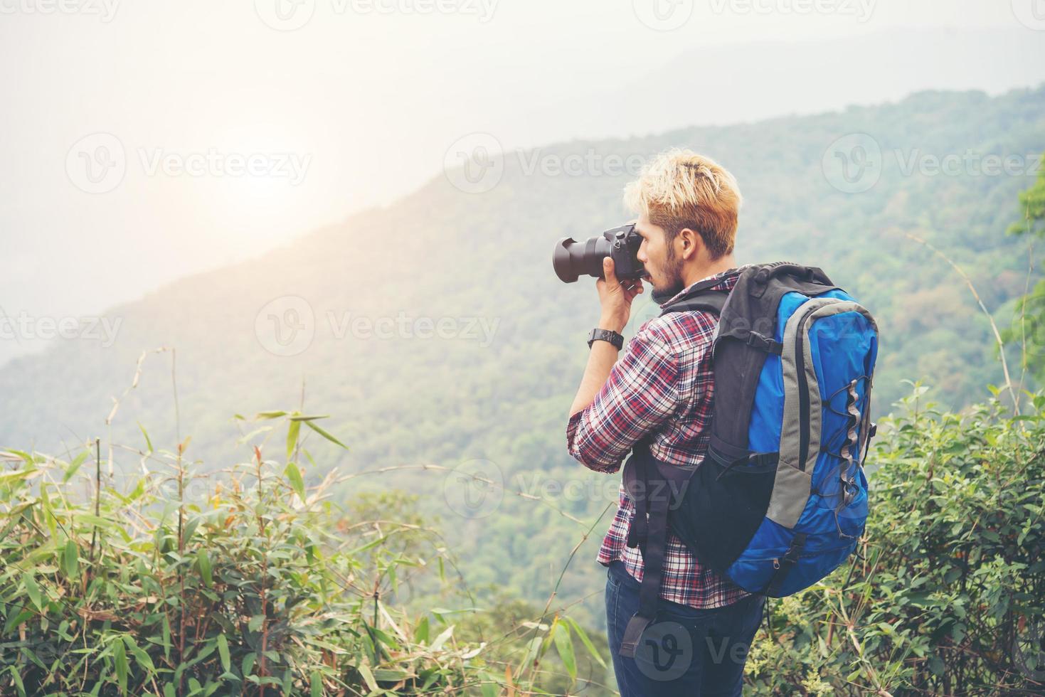 Rear of young traveller man with backpack standing on mountain and taking a photo