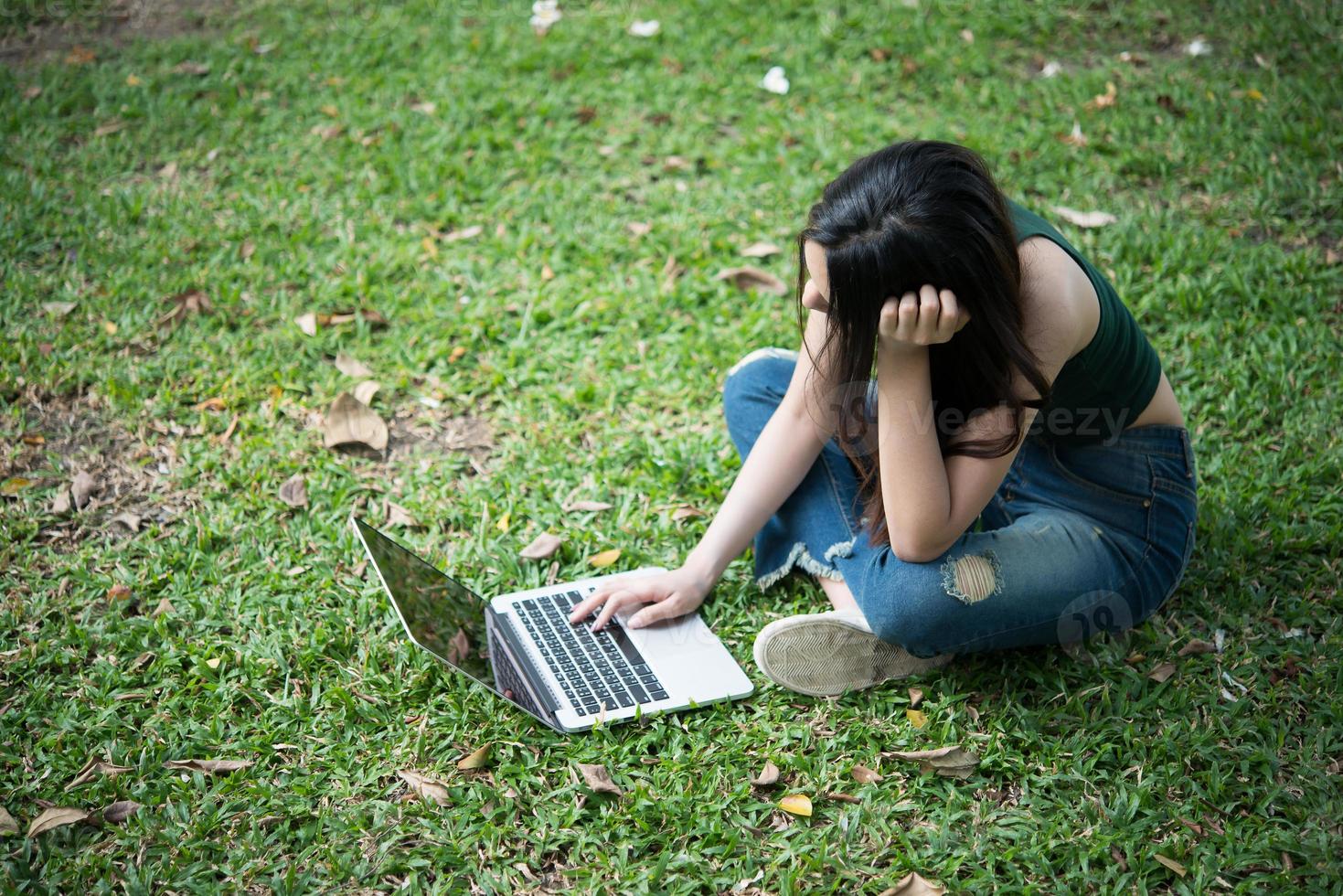 Hermosa mujer joven sentada sobre la hierba verde y usando la computadora portátil en el parque foto