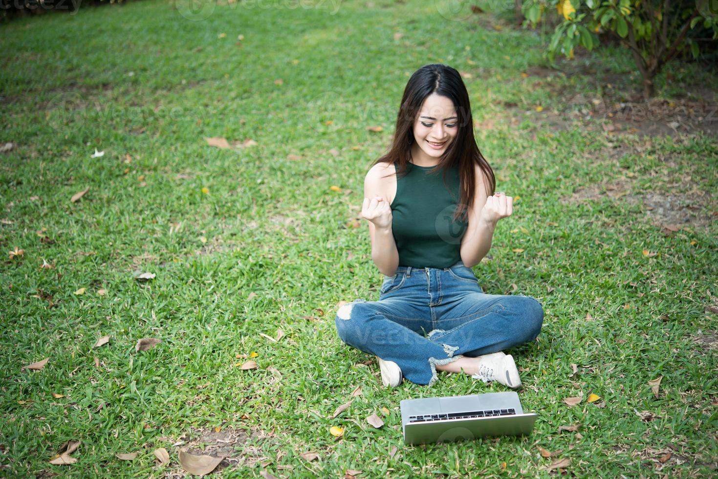 Hermosa mujer joven sentada sobre la hierba verde y usando la computadora portátil en el parque foto