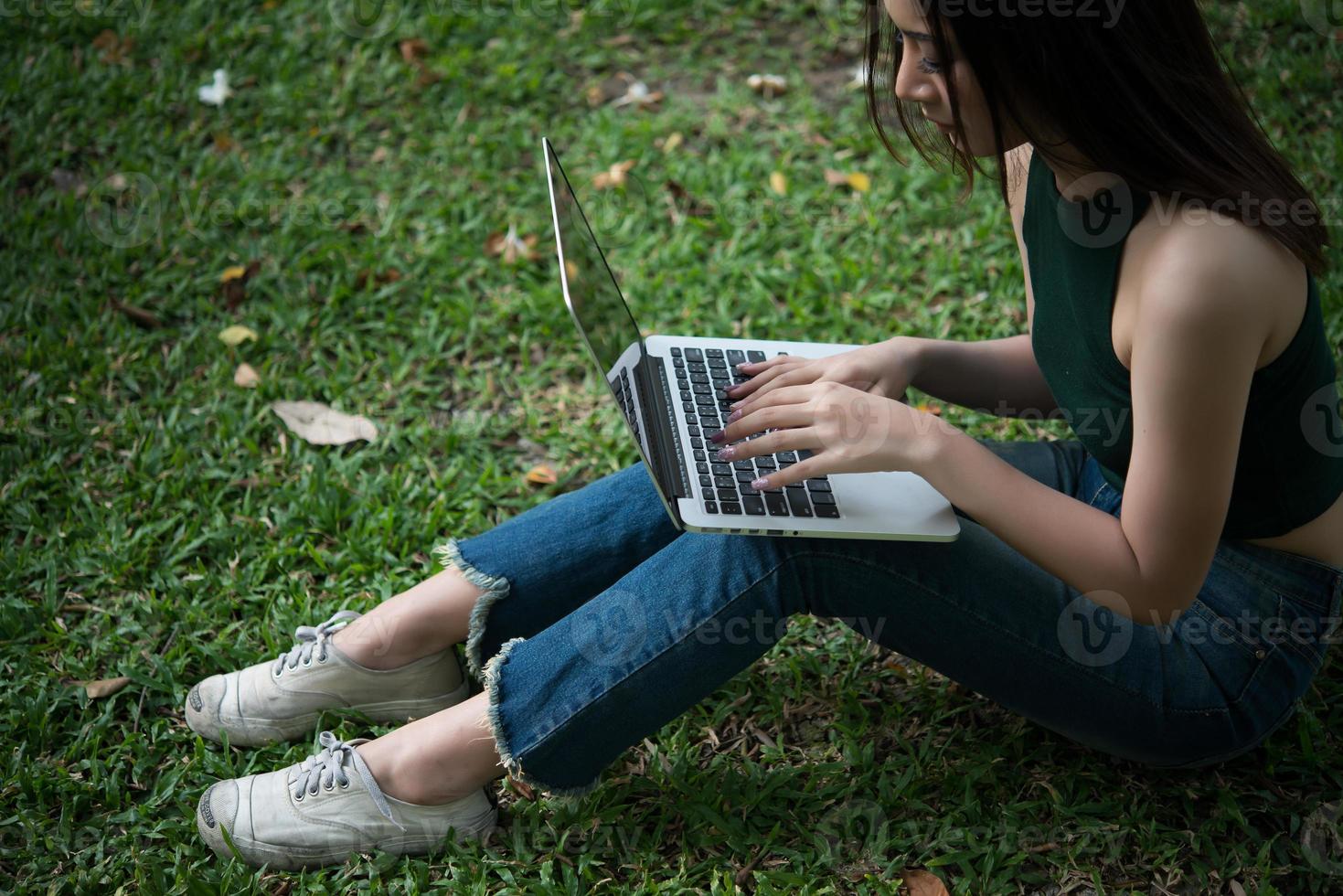 Young beautiful woman sitting on green grass and using laptop in the park photo