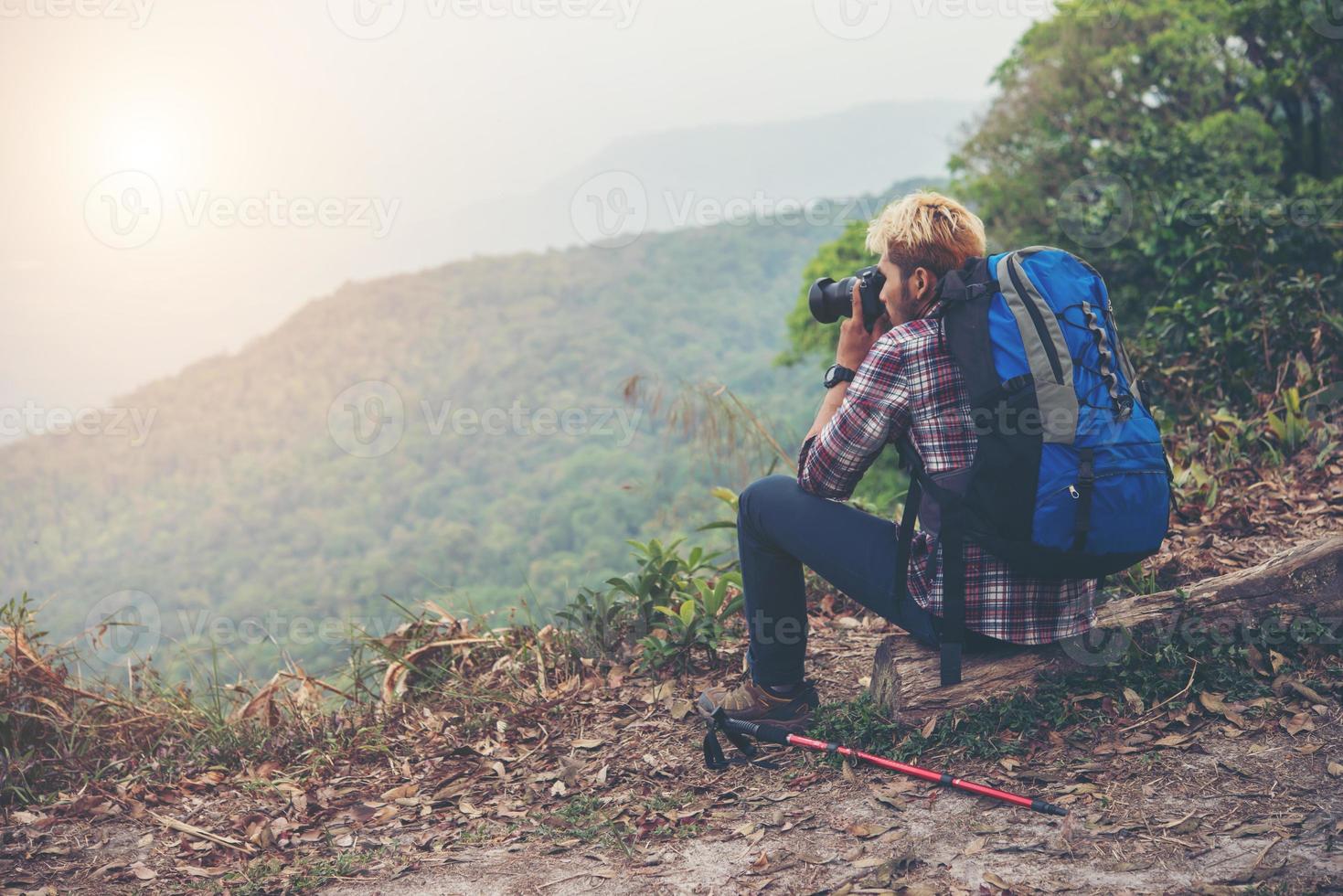 Rear of young traveller man with backpack standing on mountain and taking a photo